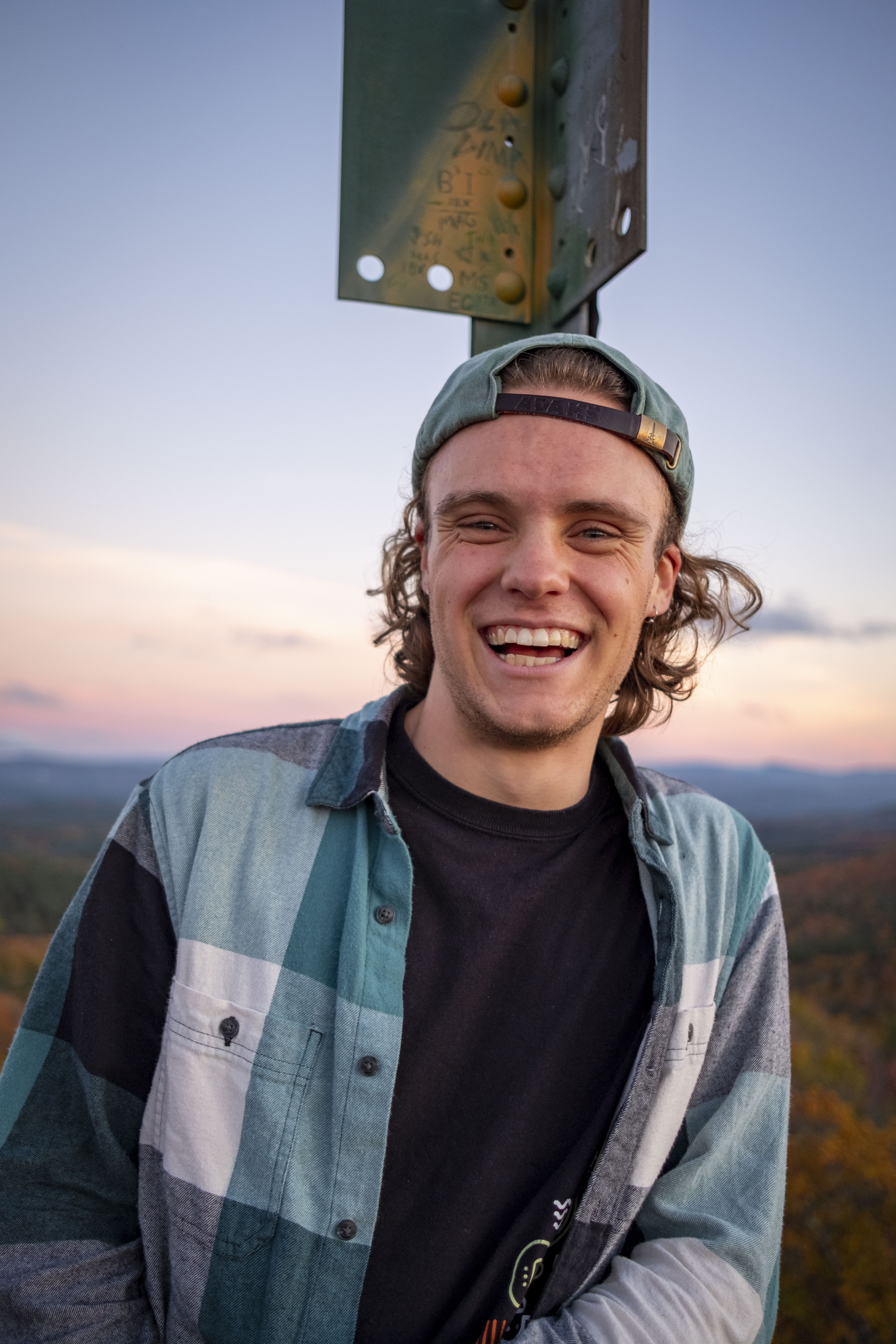 Alex standing atop the Gile mountain fire tower at sunset wearing a green flannel and a backwards baseball cap