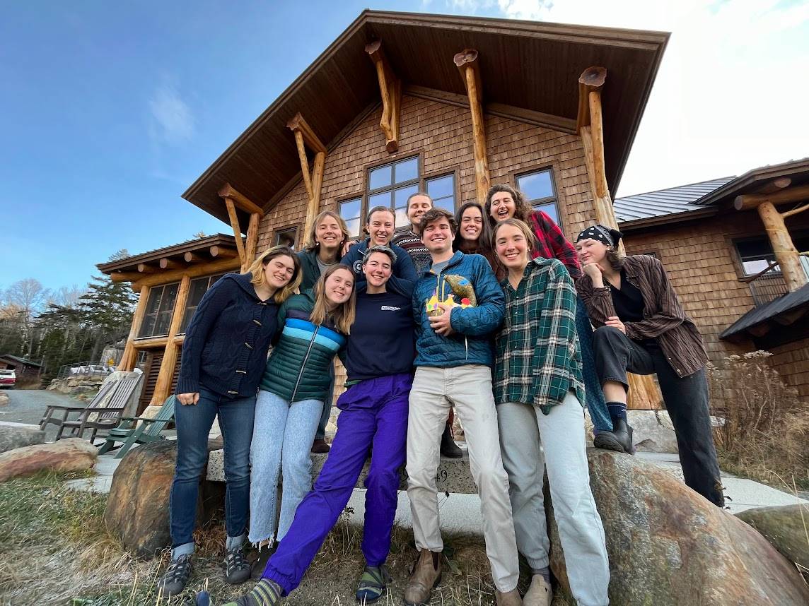 The 10 members of fall crew crew 2023 stand in front of The Lodge, a large wooden building. They're wearing coats because it is getting wintery. 